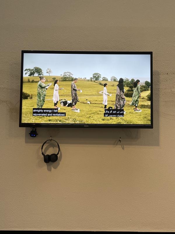 film on flatscreen. 6 figures are in a row, facing right. They are all on little step machines. They’re in a wide open rolling field with cows in the background, wearing shimmery salwars. They’re holding scripts and the wind is blowing their hair back.