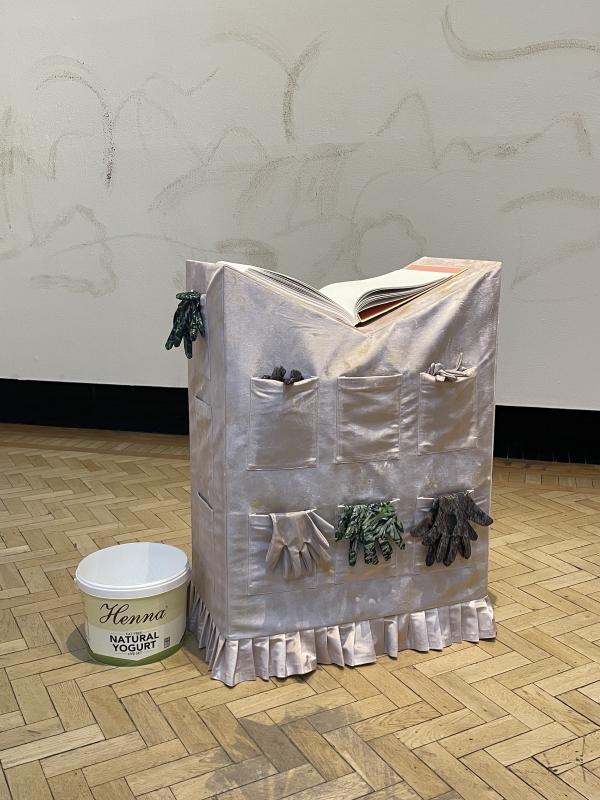 the podium with the big book. the podium is pale pink and shiny satin, there are little pockets sewn on to the side facing us, and limp little gloves hanging out of them. the bucket of henna yoghurt is on floor to the left.