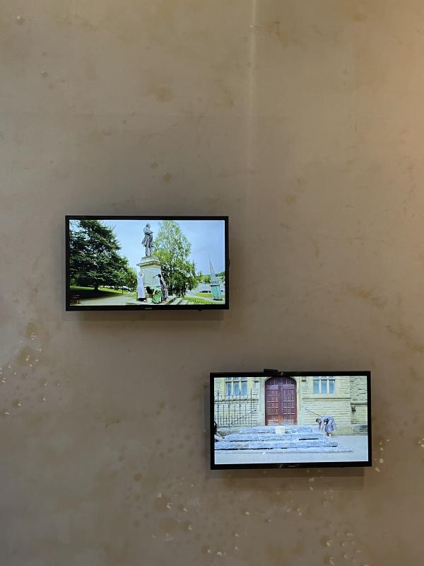 2 screens on a wall, one is higher than the other, like they're diagonal to each other. On one, the group wash the statue of John Bright with yoghurt. in the other, they wash the steps of the gallery with yoghurt. both images are too far away to see detail, mostly we can see the setting.