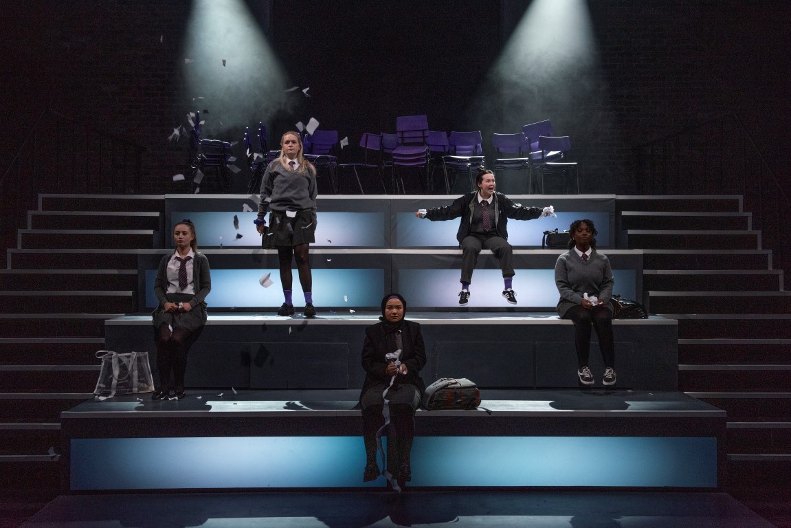 five school girls in grey and white uniforms sit on a tiered light up stage, one of them has shredded paper and is throwing it up into the air