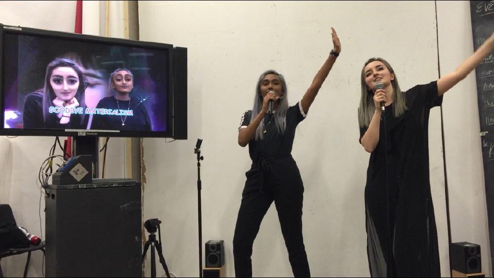 gab and zarina performing karaoke at an open studios at csm after rewriting the words to bohemian rhapsody -- the words goodbye materialism can be seen on the screen next to them