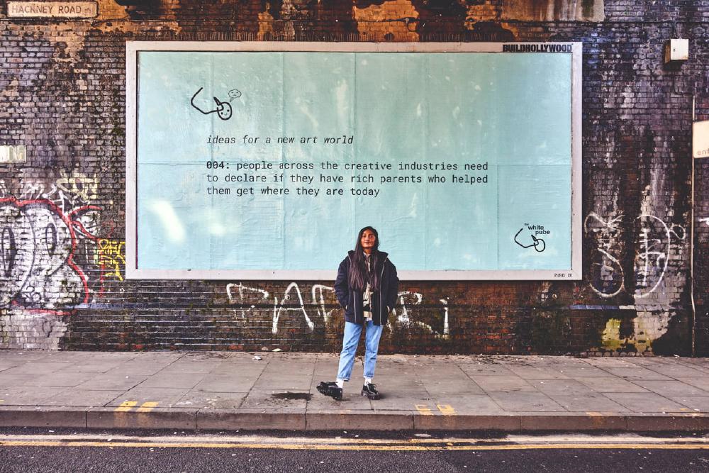 zarina standing in front of a billboard that says people across the creative industries need to declare if they have rich parents who helped them get where they are today