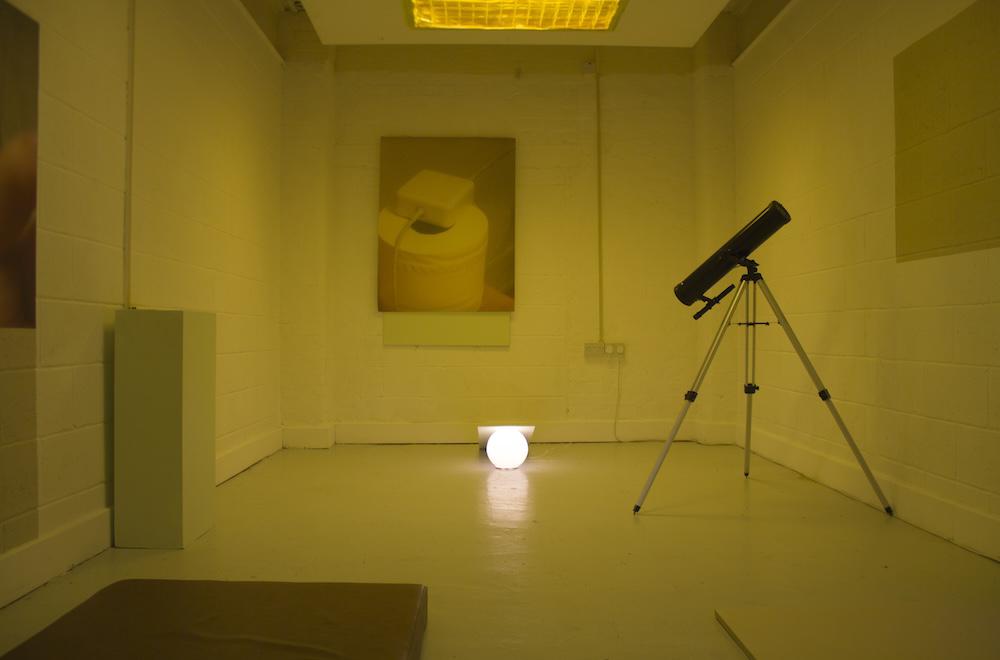 a telescope points down at a round lamp on the floor, which has wood over top to stop it shining onto the wall where there is a picture of a macbook charger on a toilet roll, and above, yellow film over the light