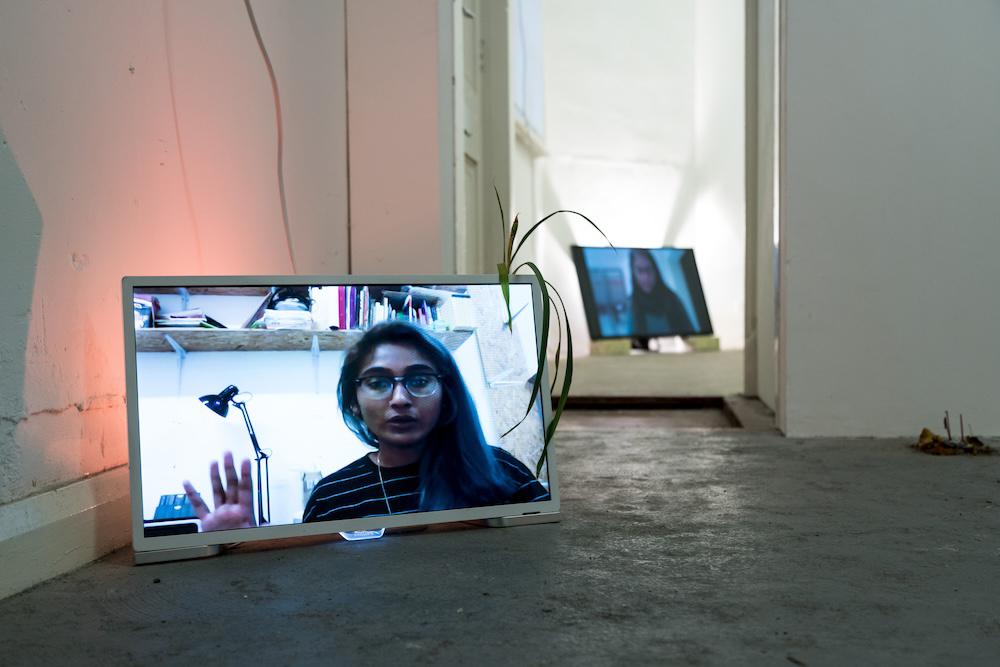 an exhibition install shot showing zarina on one screen and her mum on another behind her