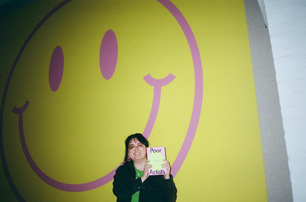 gab holding poor artists in front of a big yellow and pink smiley face on the wall that matches the colours of the book