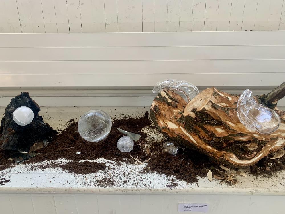 a sculpture across a flat surface is made up of a log that has been carved into to reveal different levels of wood in various tones, with mud and dirt across the bottom, and glass sculptures curving round the wood, with balls of ice appearing white against the mud