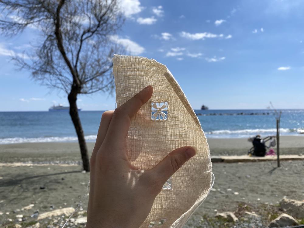 Kat holds up a pece of fabric to the ocean in front of her, the fabric is embroidered with a patterned square that has cut outs between the lines meaning you can see the ocean through the window in the fabric