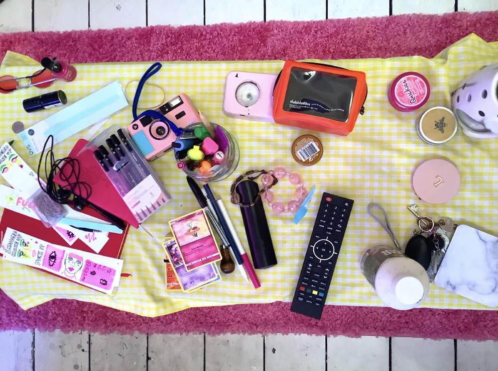 a flat lay of a table with lots of items on it: a tv remote, pink bracelet, compact mirror with the letterL on in gold, a camera, pens, highlighters, pokemon cards, sunglasses, and nail varnish