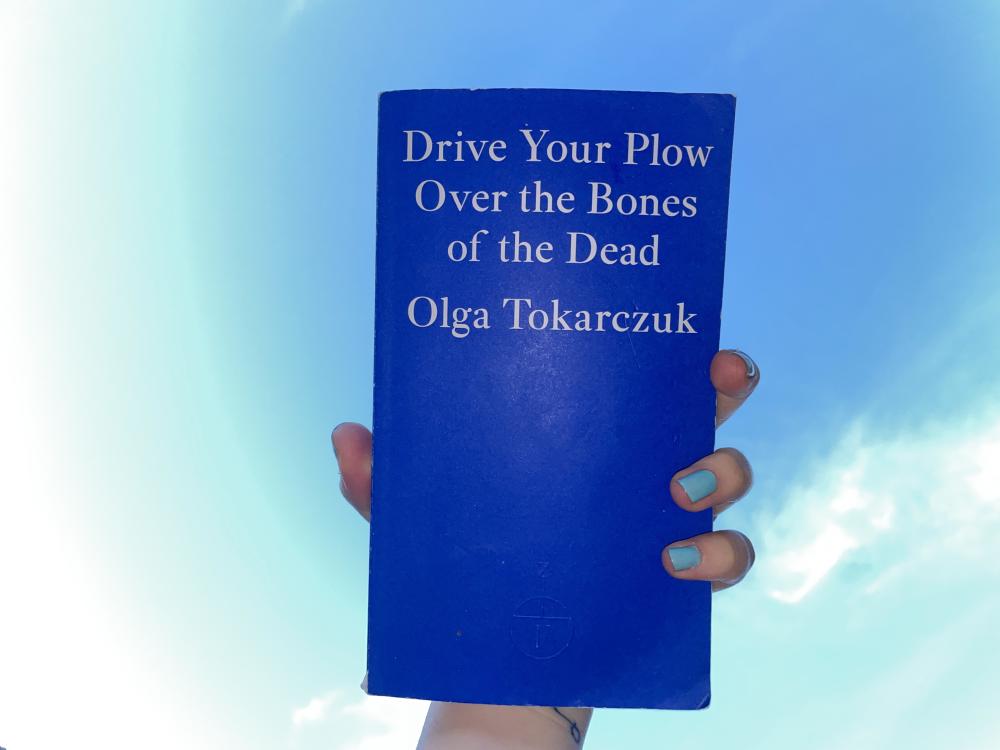 Gab holds up the dark blue Fitzcarraldo book Drive Your Plow Over the Bones of the Dead by Olga Tokarczuk against the sky, flash on, pale blue nail varnish peaking over the edge of the book