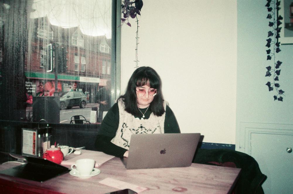 Gab in a cafe on a laptop, rain on the windows, her octagonal glasses tinted pink