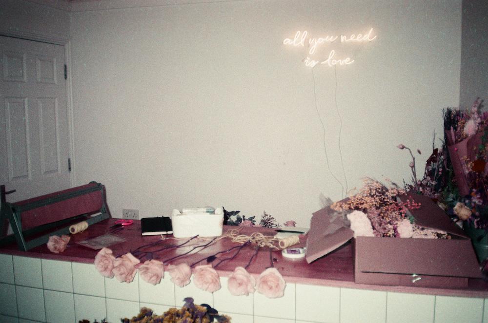 a row of pink roses on a florist's counter in front of a neon light that says all you need is love