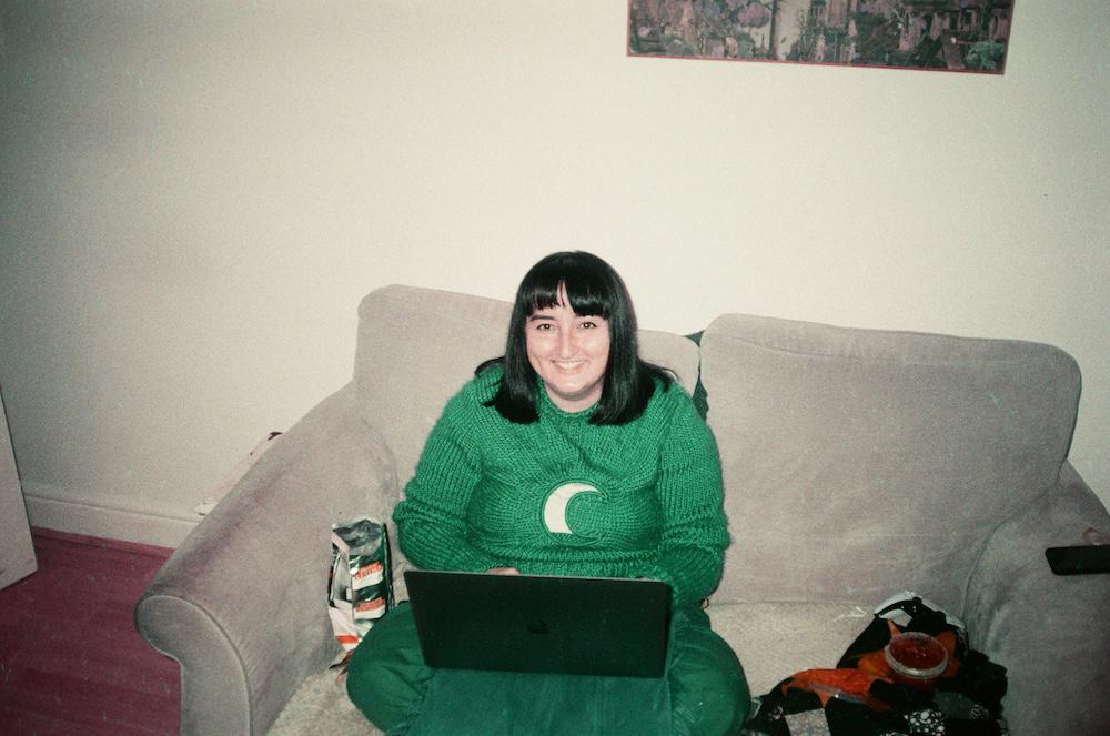 Gab smiling up at the camera with black short hair and a knitted jumper with a moon cut out of the centre, writing on a laptop on the couch