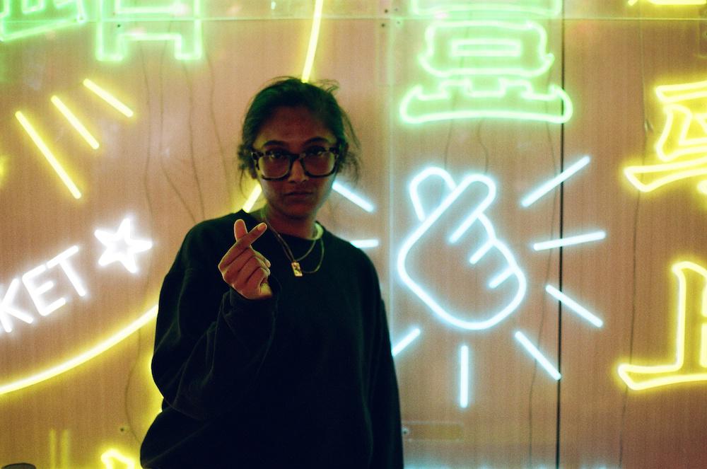 Zarina doing a heart sign with her fingers next to a neon light of the same heart-finger design