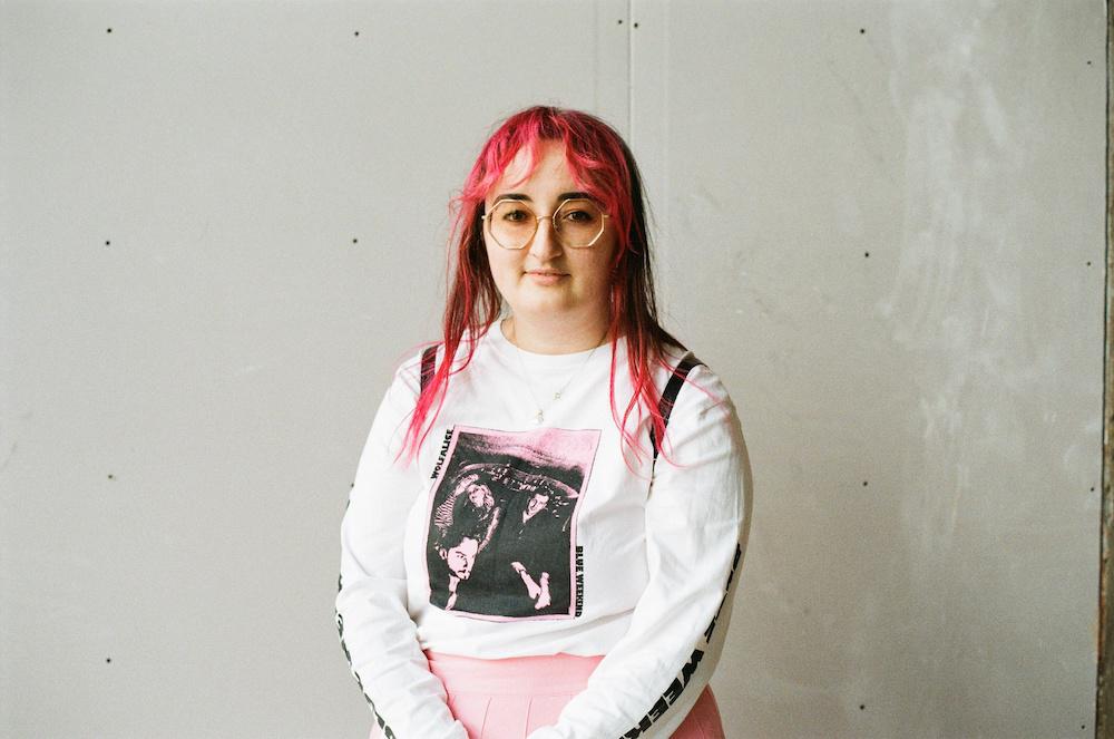 gab against a concrete wall, with pink hair and octagonal tinted glasses