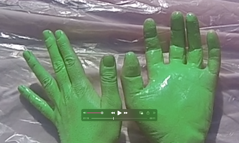 gab's hand on the left is spray painted neon green next to a fake plastic hand that is sprayed the same colour, viewed above a plastic sheet