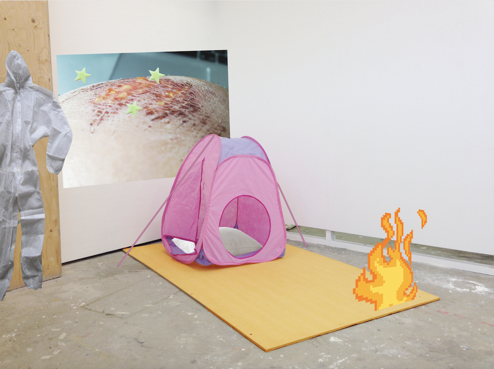 an orange board on the floor is holding a pink kids tent, and there;s a digital flame tilted to one side edited over the photograph. behind the tent there's an image of a seeping wound, and then a dust suit next to that