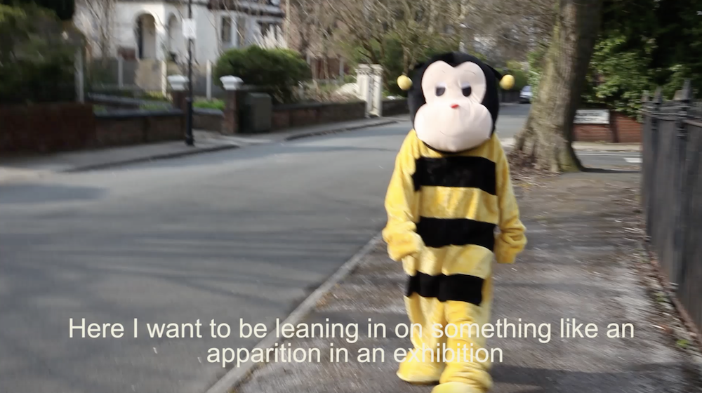 a person in a bee costume walks down a street and subtitles say here i want to be leaning on something like an apparition in an exhibition
