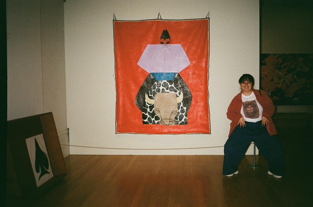 gab poses in a wide leg horse stance with hands on waist belt to match the big painting called Rodeo King next to her by Joy Yamusangie of a blocky rodeo scene painted head-on on unstretched canvas