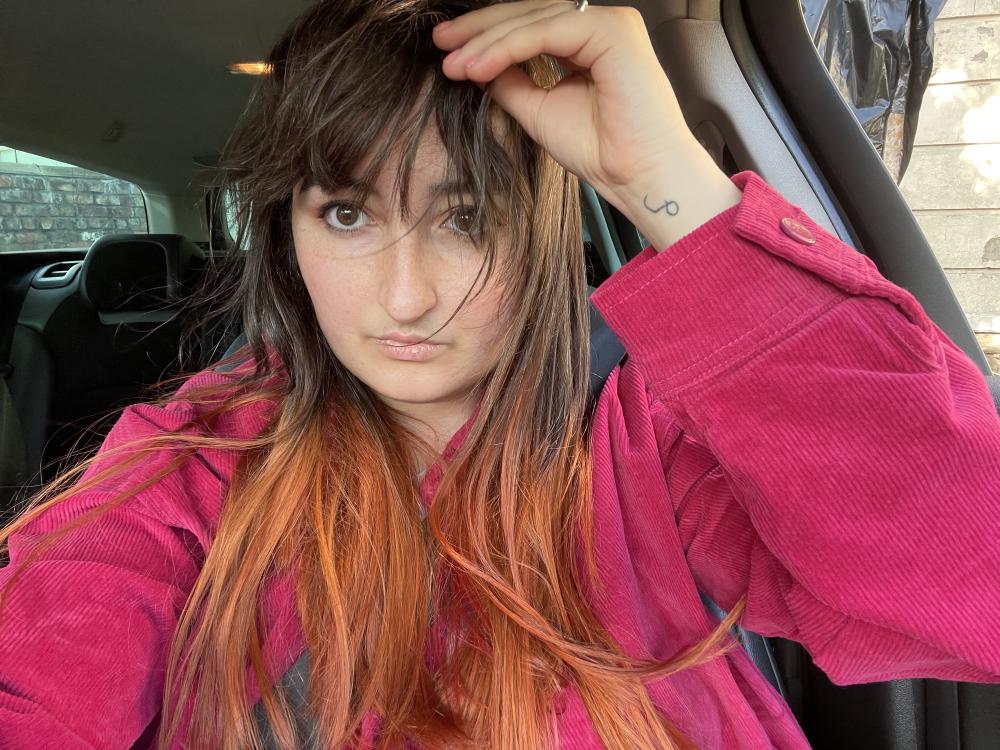 selfie in the car, the window is open so my hair was blowy; you can see my white pube logo tattoo on my wrist, and the bottom half of my hair is dyed orange