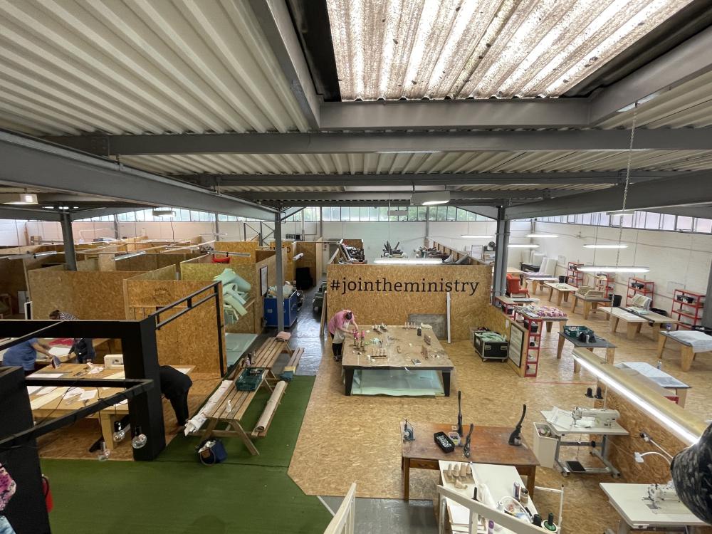 taken from the top of the stairs, you can see the whole workshop with lots of tables and machines for making upholstery and a decal on the wall that says hashtag join the ministry