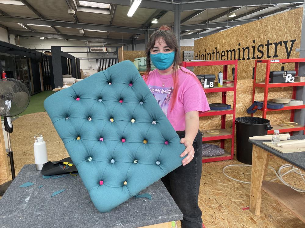 gab is holding up a deep buttoned footstool that is teal with lots of different coloured buttons, and it matches her face mask