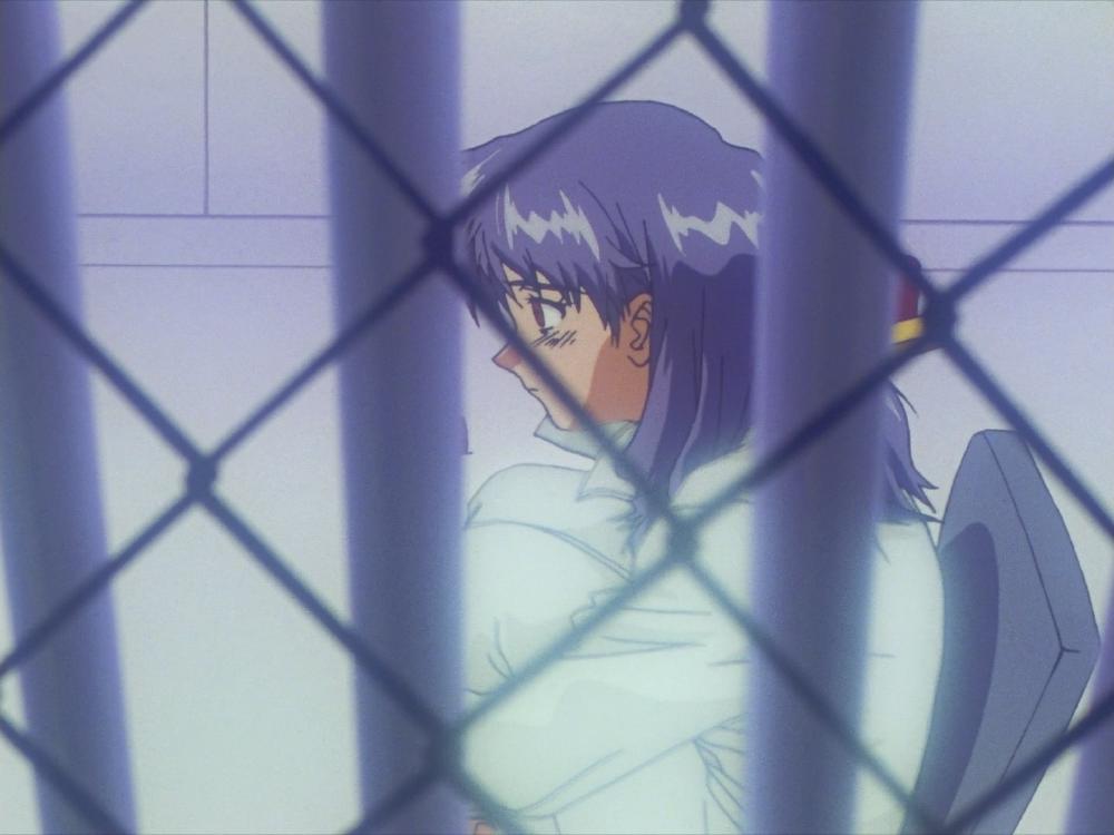 A woman sits on a chair in an asylym cell, visible through a wire fence, she looks blank