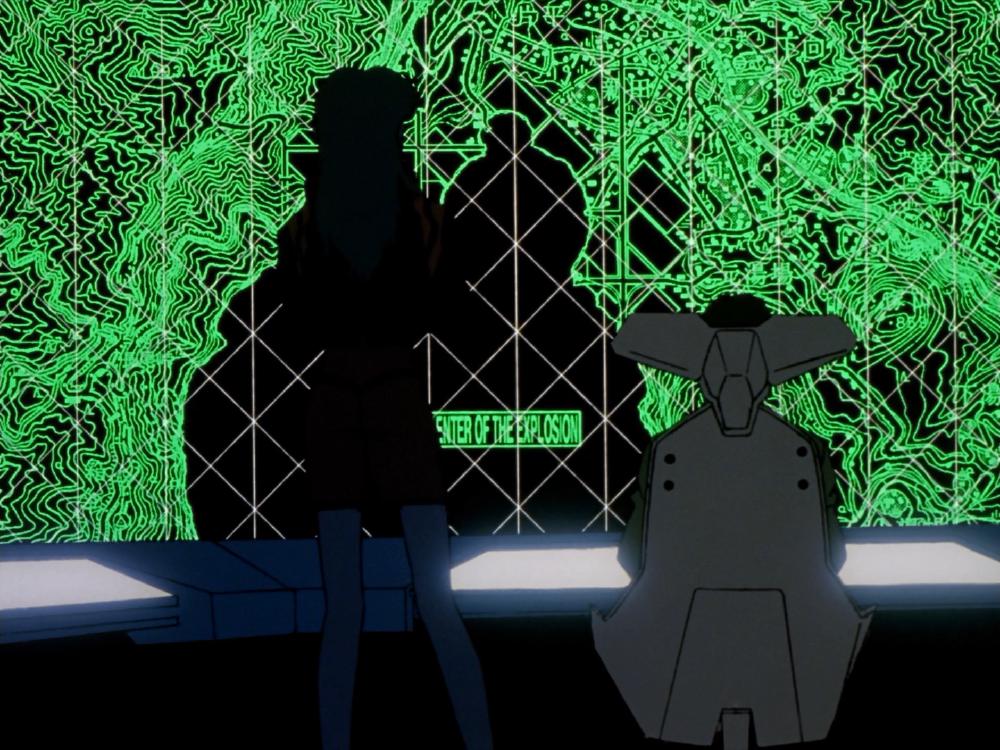 two people look at the screen in a control room, where there is a big neon green map made up of thin electric lines and topographies