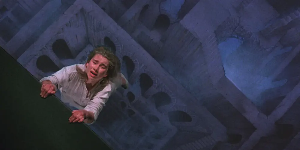 a young woman hanging off the edge of a path in hell, about to drop down into a labryinth of arches and stairways, in a screencap from hellraiser 2