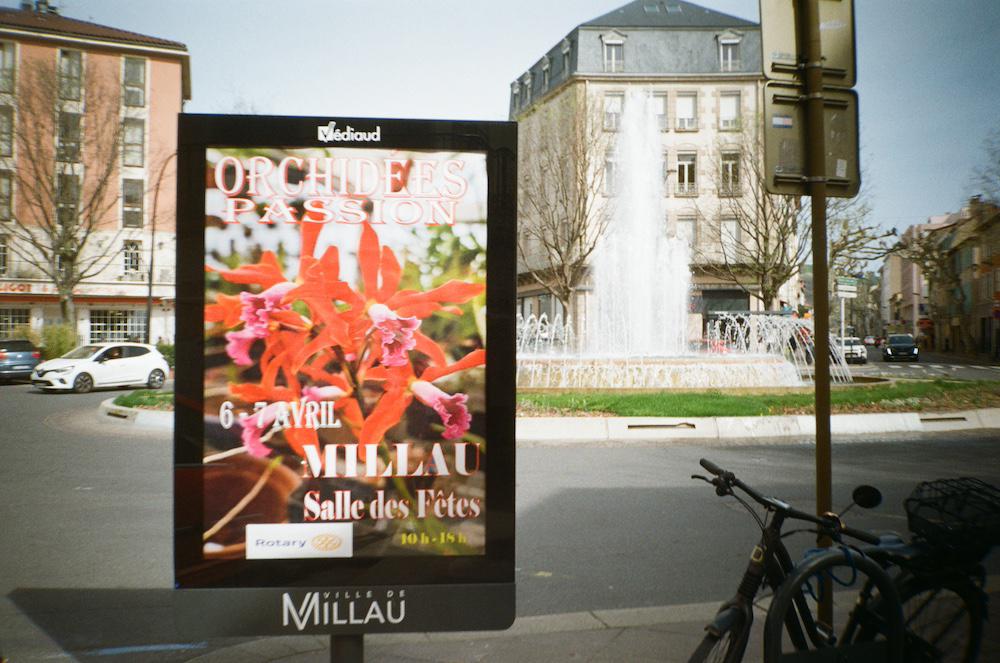 an advert for a flower festival in the centre of Millau with a fountain in the background