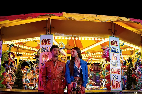 two characters stood at a fun fair against bright lights and toys at a stand