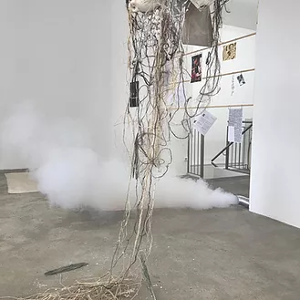 an industrial pipe on the floor spills thick white smoke onto the floor, and in the centre of the gallery there is an artwork that is like strings and nets all hanging together in a cluster