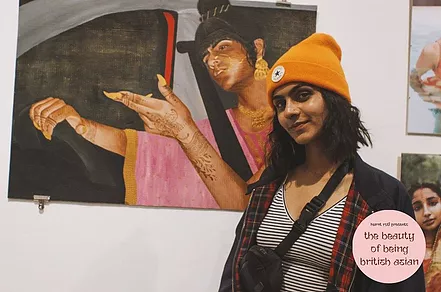 Manveer Matharu stands in front of a painting of a brown woman in the drivers seat pointing a hand out with long nails with her head titled to the side