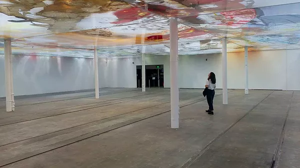 Gabrielle stands in a huge open gallery space broken up with white columns that are holding up big pieces of plastic on top of which the artist has pooled colour and texture in different shapes, so it makes a new colourful ceiling much lower than the gallery&rsquo;s original one