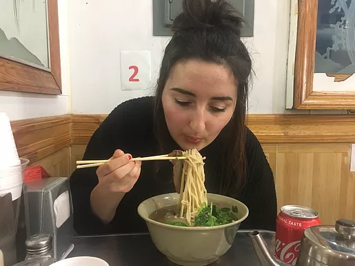 Gabrielle leans over a huge bowl of noodles blowing them before she eats