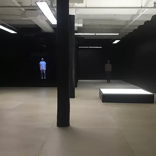 a very dark gallery space with black walls and in the distance a man stands in the space, and the same man is also projected lifesize on a video on the wall beside him