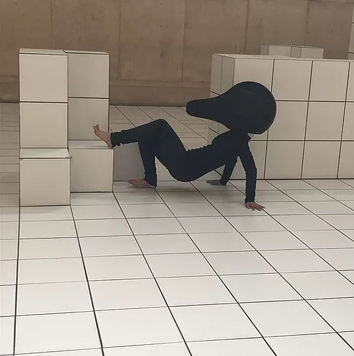 on a tiled floor with blocky sculptures that are also tiled, a performer wearing a full black skintight outfit is on all fours in a crab pose with a huge black headpiece that looks like a smooth rounded doorstopper