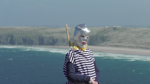 a kid stands on a cliff with a dramatic blue water and landscape behind. The kid has a silver mask all over their head, a blue and whtie striped top, and a bow and arrow on their back