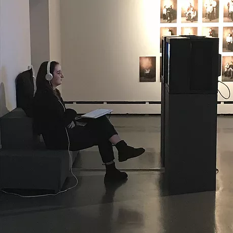yet another shot of gabrielle with her legs crossed and white headphones on watching CRTs side by side on plinths
