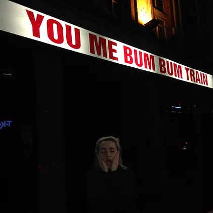 gabrielle stands outside below a red you me bum bum train sign with her hands either side of her face screaming