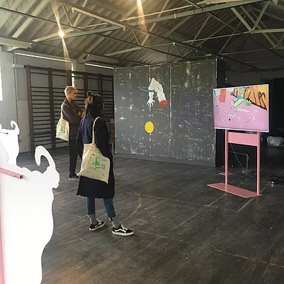 Zarina stands in the gallery watching a flat screen tv animation on a pink metal stand, next to a big painting of a white dismembered arm through a basket ball hoop pushing a yellow ball through it