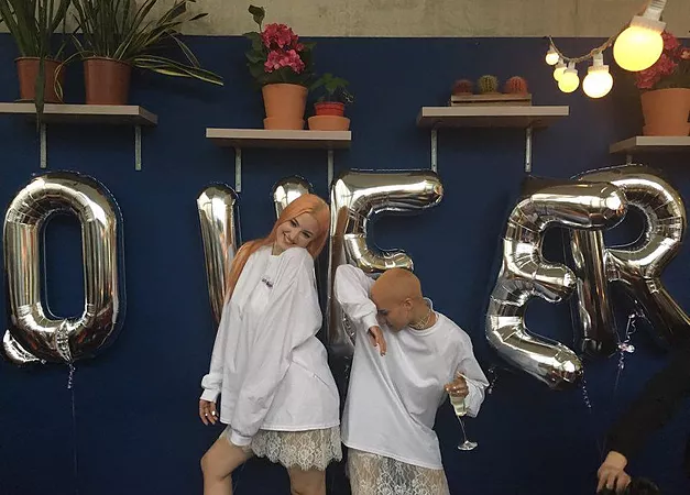 Maggie and Rene stand posting happily in their wedding outfits: all white hoodies with skirts below, behind silver bubble writing balloons that say QUEER