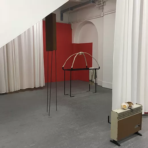 the gallery space is cut with big white curtains, and there are a few items in the space: a small electric radiator with a shell on top, and a tall metal sculpture balancing on its own feet, and another one that is shaped like a table with plaster arching on top