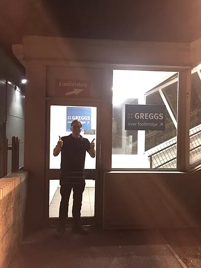GT, a white man, stands doing double thumbs up in front of a Greggs sign