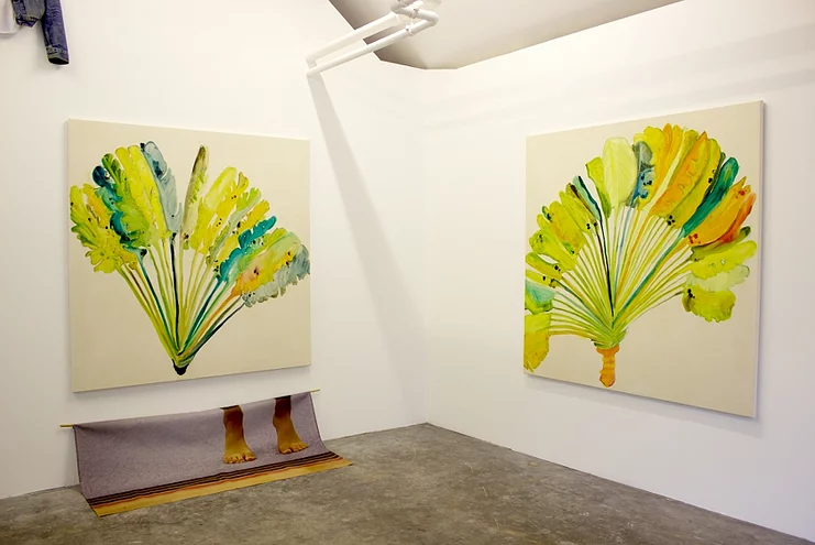 a shot of the paintings of palm fronds, and below one of them is a photographic print on a loose material of somebodys feet on tip toes