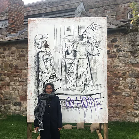 Zarina stands smiling with a big chunky scarf around her, in front of a big drawing of a native american in a headdress pointing at a british invader above the graffiti &lsquo;go home&rsquo;