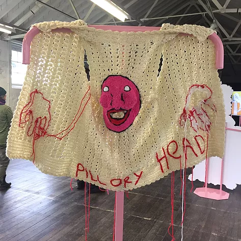 a crocheted cardigan with no arms hangs over a free standing metal rail and there&rsquo;s a scary pink face on the back of it with scary hands too and the words Pillory Head, with threads falling down too