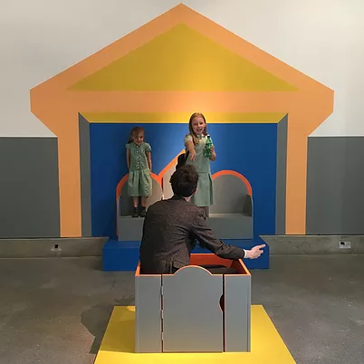 two kids point at a man sitting in a box in front of them, and there&rsquo;s a building painted on the wall behind them