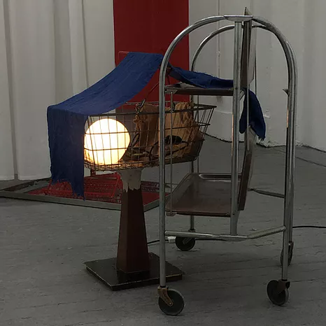 a metal trolley, next to a shopping basket being held up by a small metal plinth, with a perfect white light inside, and a strip of blue fabric draped over top