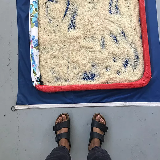 Zarina looks down at her feet in sandles in front of a big flat container of rice that looks like someone has ran their hands through