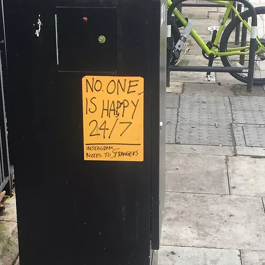 a poster on the side of an electricity box on the street says no one is happy 24 7